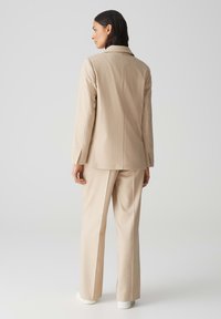 Beige op maat gemaakt pak met een gestructureerd colbert en wijde broekspijpen. De stof lijkt glad, met een klassieke notch-kraag en strakke lijnen.