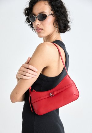 Femme aux cheveux courts et bouclés, portant des lunettes de soleil, un haut noir sans manches et un sac à bandoulière en cuir rouge avec un détail de boucle.