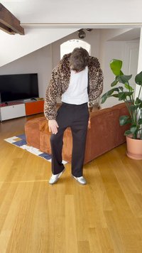 Leoparda raksta mākslīgās kažokādas jaka, balts t-krekls, melnas bikses un sudraba krāsas apavi. Koka grīda un iekštelpu augi nodrošina kontekstu.