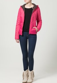 Pink quilted jacket with a brown-lined hood, front zipper, and side pockets, worn over a black top and dark blue jeans. Fringe-accented boots.