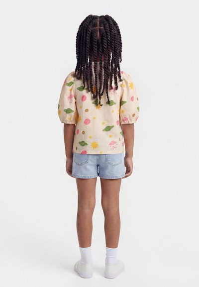 Παιδί με πλεγμένα μαλλιά που φοράει μπεζ πουκάμισο με πολύχρωμα σχέδια διαστημικού θέματος, ανοιχτόχρωμο τζιν σορτς, λευκές κάλτσες και λευκά παπούτσια, γυρισμένο από πίσω.