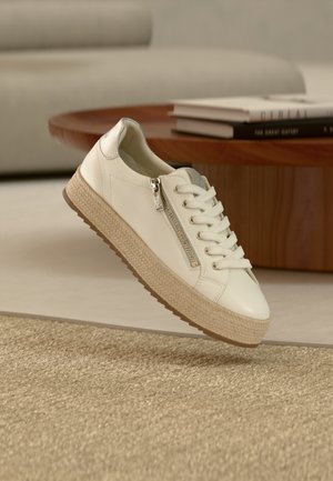 Baskets en cuir blanc avec semelle beige texturée, dotées d'une fermeture éclair latérale et de lacets, suspendues au-dessus d'un sol neutre et d'une table en bois.