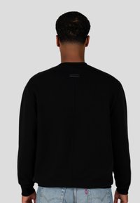 Felpa nera con colletto rotondo, polsini a costine e un logo discreto sul retro. Tessuto morbido con una texture liscia e una vestibilità rilassata.