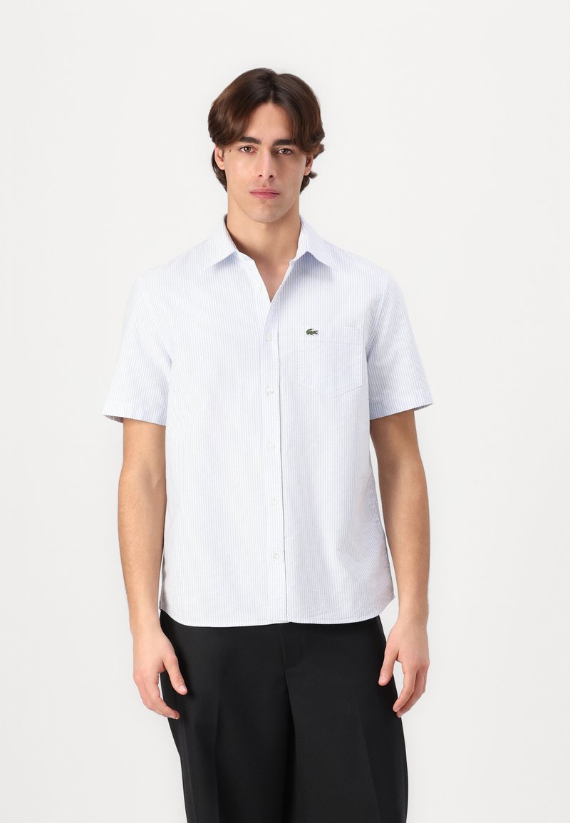 Camicia a maniche corte con bottoni di colore azzurro chiaro, con strisce verticali bianche, dotata di taschino sul petto e un logo verde sul lato sinistro.