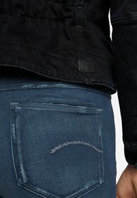 Musta tikattu farkkutakki, yhdistetty sinisiin farkkuihin, joissa on harmaa siksak-ommelu takataskussa.