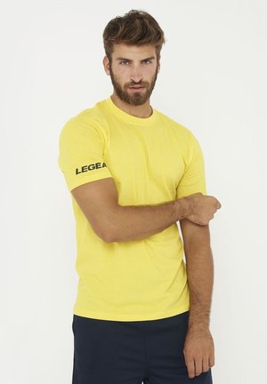 Man met baard die een geel shirt met korte mouwen draagt met de tekst "LEGEA" op de mouw, die zijn arm verstelt, staand tegen een witte achtergrond.