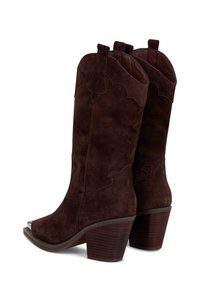 Botas de cowboy de ante marrón con punta afilada, diseño en la parte superior curvada y costuras decorativas. Presentan un tacón apilado y una puntera de metal.