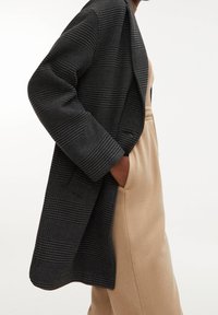 Cappotto aperto a quadri grigio scuro con spalle strutturate. Le righe verticali creano texture. Indossato sopra un outfit beige con dettagli sulle tasche.
