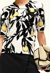 Kobieta ubrana w białą bluzkę z krótkim rękawem w czarne liściaste gałęzie i żółty wzór owoców, trzymająca beżowy pleciony kopertówkę z czarnym wykończeniem.