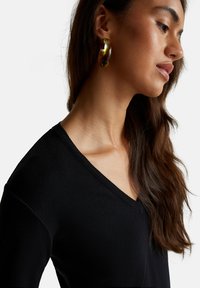Zwarte v-hals trui met een geribde halslijn. Kenmerkt een textuurstof, gestyled met grote bruine en gouden ringoorbellen. Glad haar valt neer.