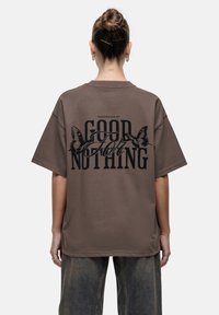 Bruine oversized t-shirt met een gedurfde zwarte graphic op de achterkant, versierd met vlinderaccenten en de tekst "PRESENTED BY GOOD FOR NOTHING."