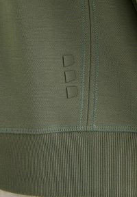 Gros plan sur un tissu vert olive avec trois ornements carrés en relief et des coutures détaillées sur une section de bord-côte côtelée.