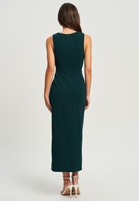 Vestido maxi sin mangas, ajustado, en verde profundo. Presenta un sutil detalle de costura en la cintura y un zipper en la parte posterior. Textura de tela suave.