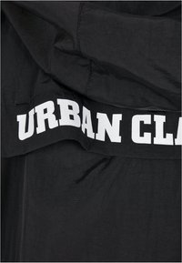 Black fabric with a smooth texture featuring a prominent white printed text reading “URBAN CLAS.” Shows a casual garment design.