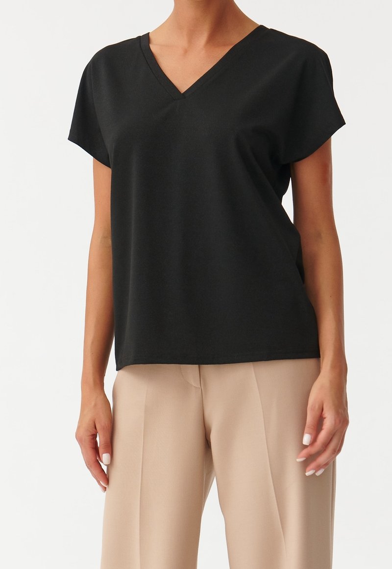 Femme portant un haut noir à manches courtes et col en V ainsi qu'un pantalon beige à jambes larges, debout devant un fond blanc uni.
