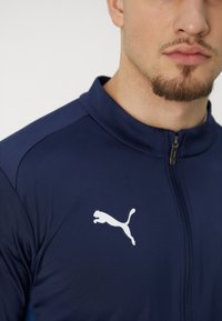 Marineblå præstationsjakke med høj krave, frontlynlås og hvid Puma-logo på venstre bryst. Glat, let struktureret stof.