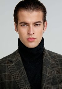 Blazer a cuadros en tonos marrón oscuro y verde, llevado sobre un cuello de tortuga negro. Tela suave con un patrón de cuadros prominente y solapas estructuradas.