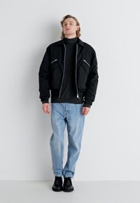 Chaqueta bomber negra con cierre frontal, dos bolsillos delanteros, puños acanalados; llevada sobre un cuello de tortuga negro, combinada con jeans de mezclilla azul claro y zapatos negros.