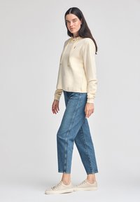 Lyse beige hættetrøje med kængurulomme, ribbede manchetter, båret med blå lige jeans og beige sneakers.