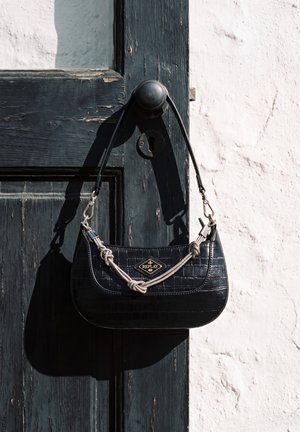 Un bolso negro de hombro Replay, con acabado texturizado y correa de cadena plateada, cuelga de una puerta de madera oscura junto a una pared blanca.
