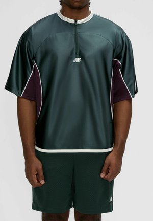 Homme portant un haut zippé à manches courtes New Balance vert foncé et un short assorti, debout devant un fond uni.