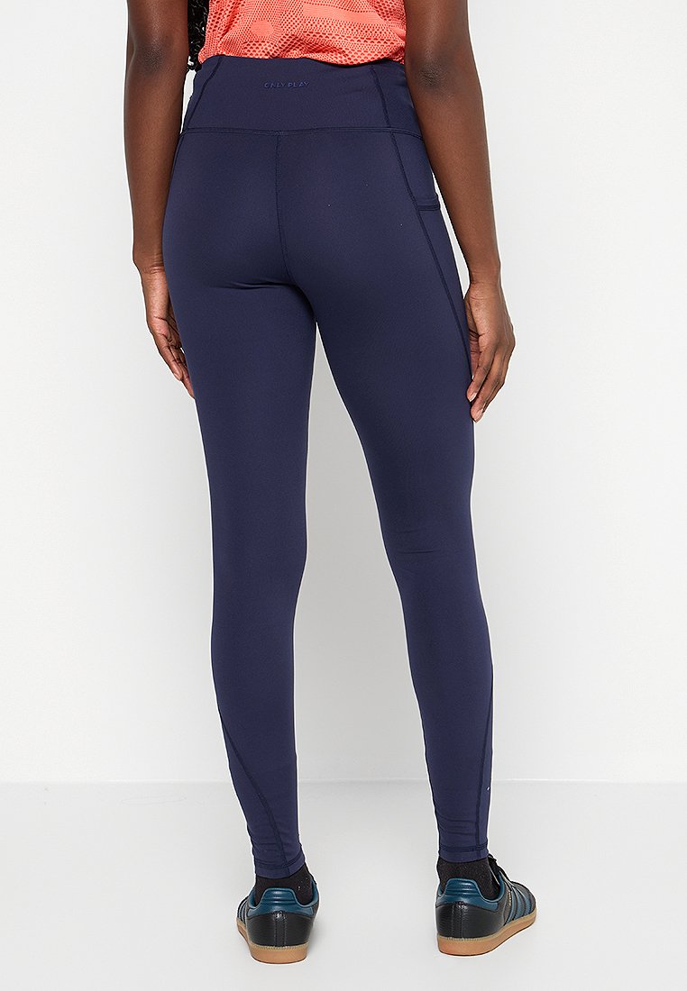 Kobieta w granatowych legginsach i czarnych butach sportowych z niebieskimi paskami, stojąca przodem do białej ściany, z rękami swobodnie opuszczonymi wzdłuż ciała.