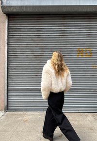 Bijela faux fur kratka jakna, široke crne hlače i smeđe cipele. Osoba hoda od metalnog roleta s hrđom i grafitima.