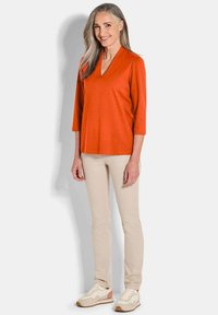 Oranje V-hals top met driekwartmouwen, gecombineerd met lichtbeige broek en witte sneakers met roze en tan accenten.