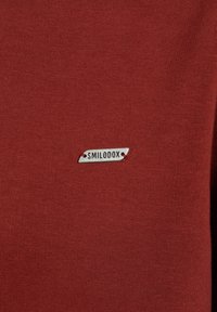 Červená bavlněná tkanina s malou stříbrnou visačkou s nápisem "SMILODOX", připevněnou červenou nití. Hladký povrch.