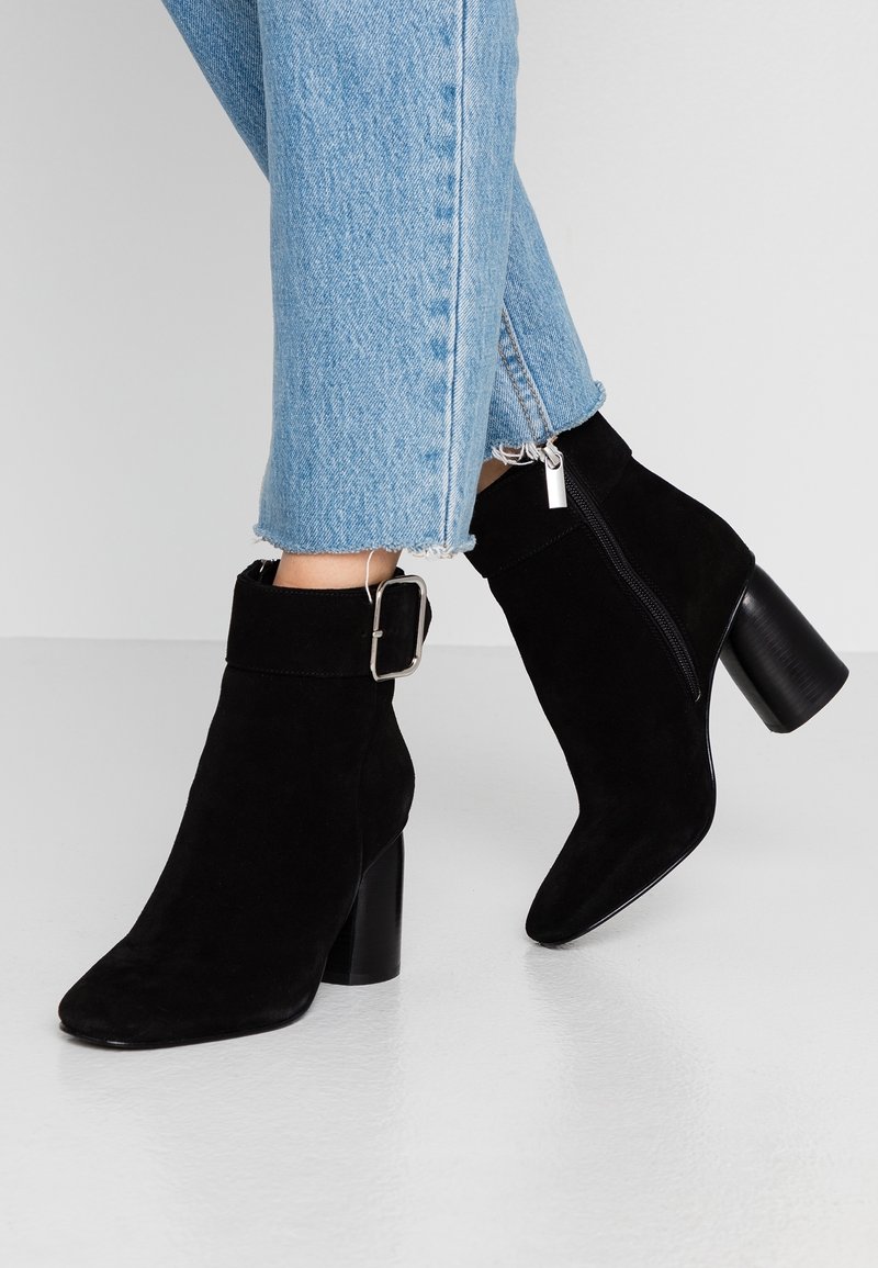 Bottes cheville en daim noir avec un talon carré, un bout pointu, et une bride de cheville ornée d'une boucle argentée. Fermeture éclair visible sur le côté.