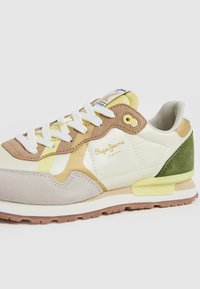 Zapatilla multicolor con gamuza beige, cuero blanco, acentos amarillos y panel trasero verde. Cordones gruesos blancos; suela de goma con tracción.