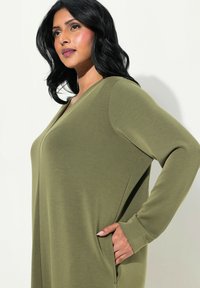 Tunique verde oliva a maniche lunghe con scollo a V e tasche laterali, realizzata in tessuto morbido con una vestibilità rilassata. Maniche e cuciture testurizzate visibili.