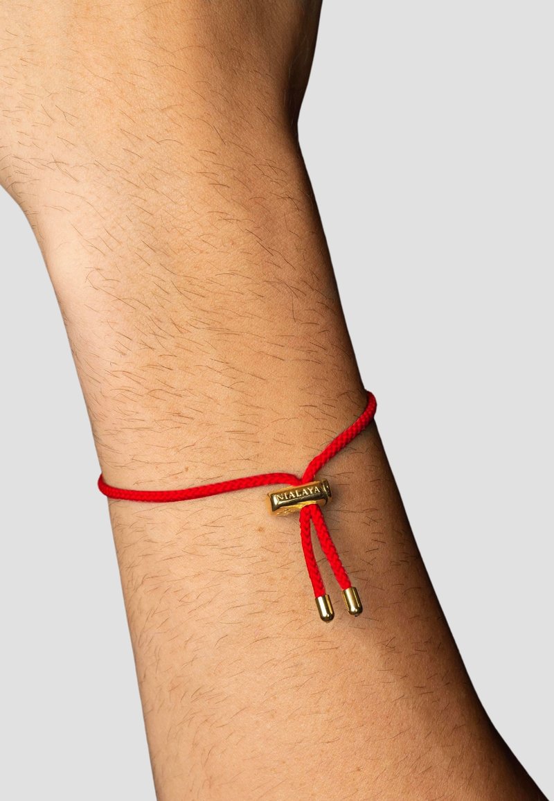 Rød snorarmbånd med et guldaccentstykke og to guldspidsede ender, hviler på et håndled og viser en glat hudstruktur.