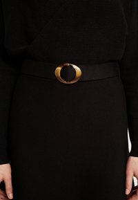 Robe noire en tricot côtelé avec manches longues et boucle de ceinture ovale en écaille de tortue à la taille, mains visibles de chaque côté.