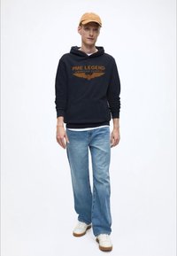 Temno modra jopa s kapuco z oranžnim logotipom in grafiko kril, modre kavbojke in bele superge z rjavimi podplati, dopolnjene s svetlo rjavo kapo.