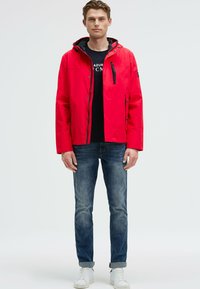 Veste imperméable rouge avec une capuche, poche zippée sur la poitrine, fermeture à bouton et poches latérales. Portée par-dessus une chemise noire et un jean bleu.