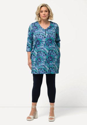 Tunica a motivi blu e verdi con scollatura a V e maniche tre quarti, abbinata a leggings scuri e sandali a tacco basso chiaro.