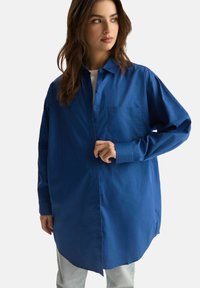 Koszula z długim rękawem w intensywnym niebieskim kolorze, wykonana z bawełny. Posiada kołnierzyk zapinany na guziki, kieszeń na przodzie oraz zakrzywiony hem dla swobodnego kroju.