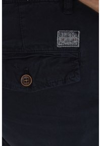 Pantalones azul marino de tela suave con cierre de botón frontal, que presentan una etiqueta de parche y detalles de costura sutiles cerca del bolsillo.