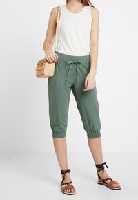 Pantalones verdes cortos con puños elásticos, combinados con una camiseta sin mangas blanca y sandalias beige. Un pequeño bolso de madera completa el conjunto.