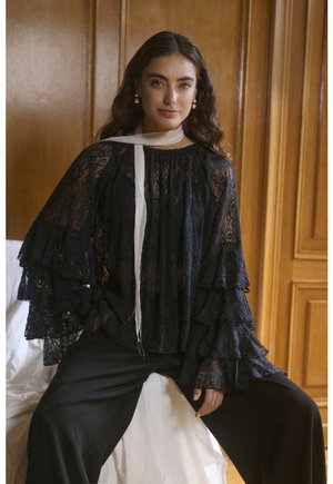 Crna čipkasta bluzica s volanima na rukavima, s bijelim šalom. Model sjedi, nosi crne hlače. Drvena pozadina dodaje toplinu.