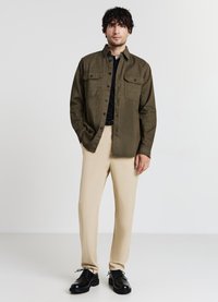 Camicia verde oliva con bottoni, dotata di due tasche sul petto, abbinata a pantaloni beige e scarpe nere stringate, su uno sfondo neutro.