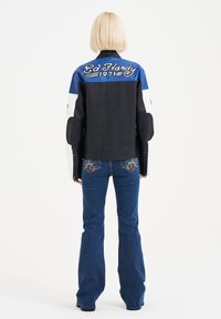 Giacca da motociclista in pelle nera con dettagli blu e bianchi, con ricamo "Ed Hardy 1971" sulla schiena. Indossata con jeans a zampa.