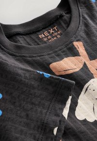 Gros plan sur un t-shirt noir pour enfants avec un imprimé de chien de dessin animé, des empreintes de pattes bleues sur la manche, et une étiquette intérieure portant l'inscription "NEXT LOVE ME KEEP ME PASS ME ON."