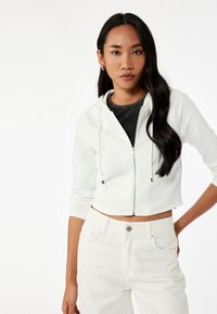 Witte cropped hoodie met een rits aan de voorkant en een trekkoord, gecombineerd met witte high-waisted jeans. Glad textuur en minimalistisch ontwerp.