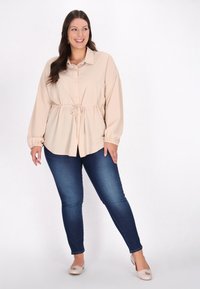 Lys beige button-up skjorte med snoretalje, parret med mørkeblå skinny jeans og neutrale flade sko, sat mod en hvid baggrund.