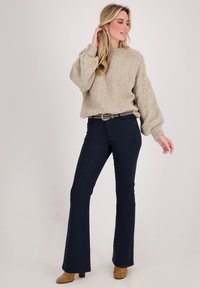 Beige grove gebreide trui met volumineuze mouwen, gecombineerd met marineblauwe uitlopende jeans, geaccentueerd met een ceintuur met patroon en tan enkellaarsjes met hak.