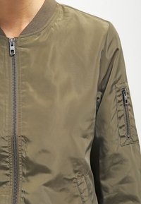 Olivengroen bomberjack van soepel nylon, met een rits aan de voorkant, geribbeld kraag en een ritszak op de linkermouw.