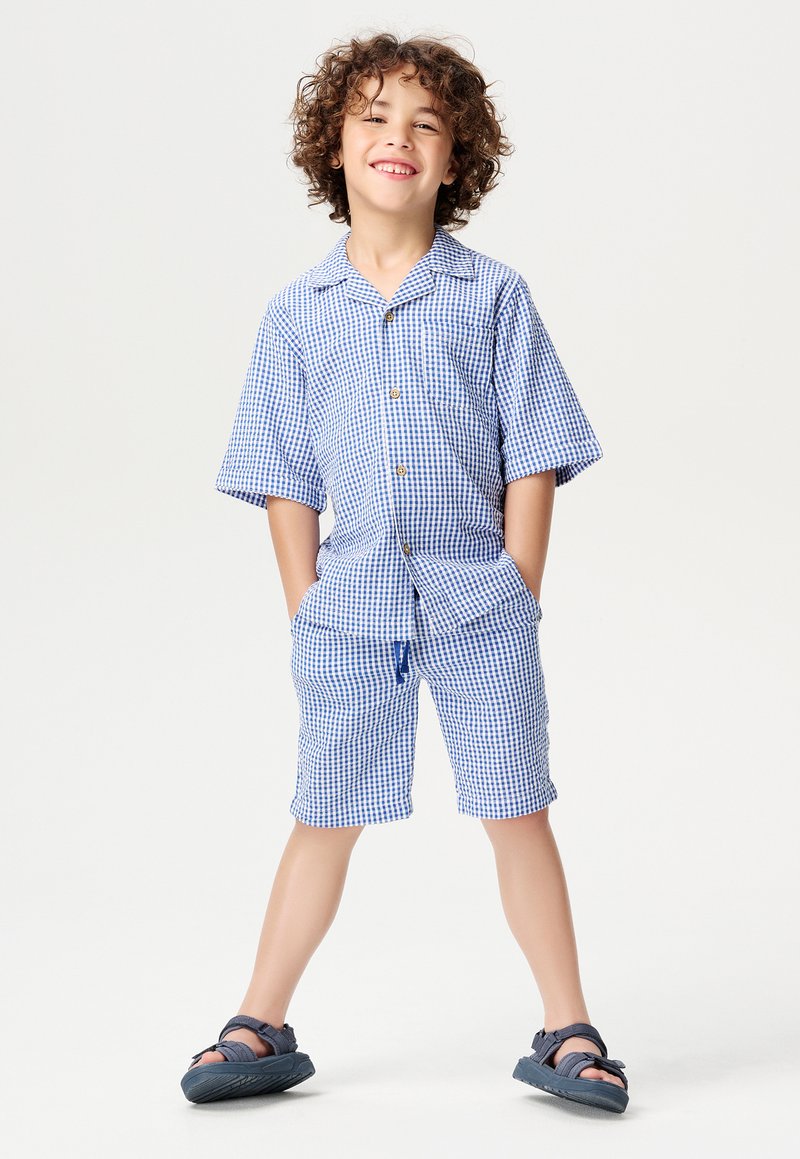 Ensemble de chemise à manches courtes et de shorts à carreaux bleus et blancs avec des boutons, comprenant des poches avant. Porté avec des sandales navy.