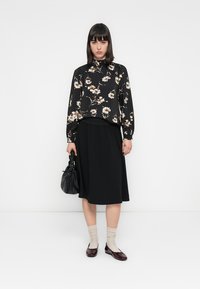 Svart blomstret bluse med lange ermer, paret med et svart, knelangt skjørt. Tilbehør inkluderer en teksturert svart håndveske og burgunder loafers.